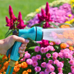 Teal Sprout garden nozzle in hand emitting a steady soaker-style spray over pink and orange bedding plants