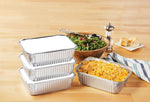 Stacked aluminum takeout pans with folded lids beside a filled pan of mac and cheese, salad bowl and bread on a table.