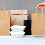 Two lidded Handi-foil aluminum meal prep pans stacked in front of brown and black paper bags on a gray backdrop.
