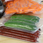 Stack of vacuum-sealed bags containing carrots, cucumbers and sausage links on a countertop