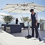 Outdoor terrace scene with a staff member sweeping and using a black upright dustpan under a large patio umbrella.
