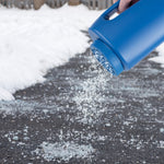 Blue handheld spreader pouring salt or ice melt onto a snowy driveway from its perforated lid