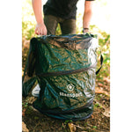 Outdoor lifestyle image of a person placing or adjusting a green Stansport trash can with a black liner in a wooded setting.