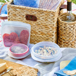 Picnic scene with Stasher reusable bags and silicone stretch lids holding snacks beside a woven picnic basket