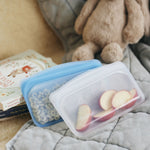 Two Stasher silicone bags, one blue and one clear, filled with cereal and sliced fruit on a quilted surface