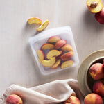 Clear Stasher silicone bag filled with peach slices on a light tabletop with whole peaches and a bowl nearby