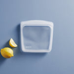 Single clear square Stasher silicone bag on a blue surface with a lemon and lemon wedge nearby