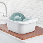 White Sterilite dishpan holding soapy water and green plates, shown on a wooden board beside a kitchen sink