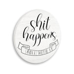 Round white wood-grain wall plaque with black script “shit happens” and a ribbon reading “roll with it” illustration