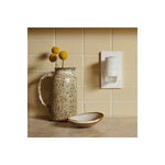 Styled kitchen countertop scene showing a plugged-in white oil warmer beside a ceramic pitcher and dish