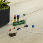 Styled scene of test tubes in a green rack, loose colored capsules, pipette and small piles of soil