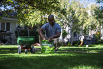 Wide lawn scene with person kneeling and placing a Sunday Green Machine bag on the grass beside a push spreader