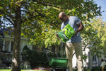 Person pouring Sunday Green Machine granular fertilizer from a bag into a green lawn spreader under a large tree
