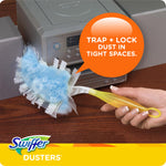 Hand holding a Swiffer duster cleaning near electronics with blue duster head and orange callout reading 'Trap + Lock dust in tight spaces.'