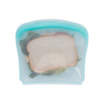 Top-down view of a teal reusable silicone sandwich bag holding a lettuce and cheese sandwich on white background