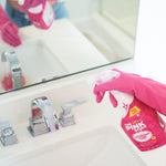 Gloved hand spraying The Pink Stuff bathroom foam cleaner toward a bathroom sink and mirror, reflection visible in the mirror