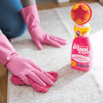 The Pink Stuff bottle with star brush standing on a rug while a person wearing pink gloves wipes the surface with a cloth