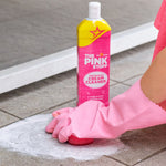 Pink-gloved hand scrubbing outdoor tile with a sponge while a bottle of The Pink Stuff cream cleaner stands nearby