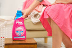 Lifestyle scene of a person in a pink dress placing The Pink Stuff Miracle Laundry bottle on a wooden stool near laundry basket.
