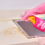 Gloved hand spraying The Pink Stuff multi-purpose cleaner at a stained countertop edge next to a stainless sink