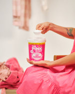 Close-up lifestyle photo of hands scooping powder above an open tub of The Pink Stuff Oxi Powder in a laundry setting