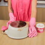 Person in pink gloves and dress demonstrating cleaning a heavily soiled pan with The Pink Stuff tub visible on the table.