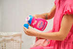 Person in a pink dress pouring The Pink Stuff Miracle Laundry Sensitive detergent into the bottle cap beside a laundry basket.