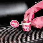 Pink-gloved hands using a sponge to scoop The Pink Stuff from an open tub placed on a grill grate.