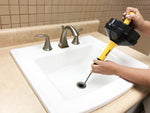 Hands operating a yellow and black TheWorks manual drain snake inserted into a bathroom sink drain
