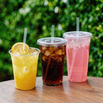 Three clear disposable cups with lids and straws filled with yellow, dark cola and pink beverages on a wood surface outdoors