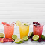 Three colorful cocktails served in clear plastic cups on a marble surface, garnished with lime, grapefruit and cranberries.