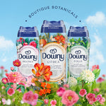 Three Downy Boutique Botanicals scent booster bottles (Peonia, Citrea, Folia) displayed among colorful flowers on a sky background.