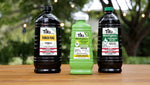 Three TIKI torch fuel containers on a wooden table outdoors: two dark bottles flanking a green clean-burn formula bottle, blurred background