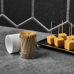 Clear dispenser of bamboo toothpicks and white ribbed cap next to a plate of cheese cubes on a dark textured surface