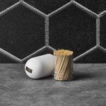Styled photo of the white ribbed cap and clear dispenser of natural bamboo toothpicks against a textured hexagonal backdrop