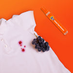 White shirt with red stain and grapes beside a Tide To Go OXI pen on an orange background suggesting spot-treatment use