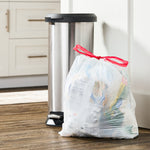 Kitchen lifestyle photo showing a tied white drawstring garbage bag next to a stainless step trash can on a wood floor