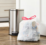 White drawstring trash bag tied with red ribbon sitting on floor next to a stainless step trash can in a home setting