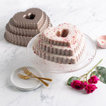 Bronze tiered heart bundt pan next to a frosted multi-tier heart cake on a glass stand with roses and forks