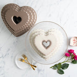 Bronze tiered heart bundt pan beside a glazed heart-shaped cake on a glass cake stand with forks and roses