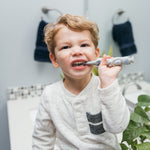 Toddler brushing front teeth with a gray otter-shaped toothbrush in a bathroom