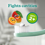 Close-up of a toothbrush with a dollop of white toothpaste and illustrated orange and mango characters with an ages 2+ badge.