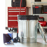 Lifestyle scene with a metal trash can lined with a black drawstring bag, a box of Great Value Strong Flex bags and a broom in a garage setting.