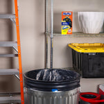 Metal trash can lined with a black Hefty bag in a garage scene with a Hefty product box displayed on a wire shelf behind it.