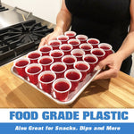 Person holding a metal tray filled with many red mini plastic cups containing red gelatin or liquid, photographed in a kitchen