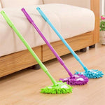Three triangular mops in green, purple, and blue leaning against a sofa on a wood floor, showing full handles and mop heads.
