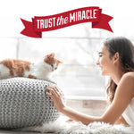 Lifestyle image of a smiling woman reaching toward a cat on a knitted pouf under a 'Trust the Miracle' banner