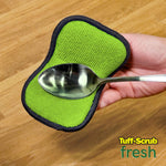 Hand holding a green dual-sided microfiber scrub pad with a spoon placed on top, wood surface background and small Tuff-Scrub Fresh logo