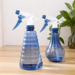 Two blue refillable trigger spray bottles in different ribbed and teardrop shapes placed on a wooden table with plants in the background.