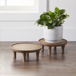 Lifestyle image of two low round wooden plant stands on a wood floor, one holding a white planter with green foliage